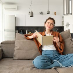 Portrait of young smiling beautiful woman, sitting on sofa in her living room, holding study material, doing homework, reading notes in notebook.
